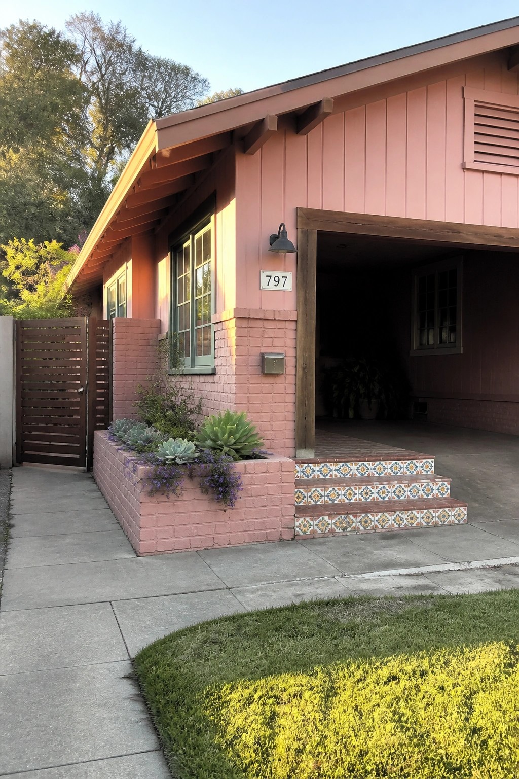 A single-story pink bungalow exterior with brick base, tiled entry steps, wood trim, and a raised planter bed beside the driveway.
