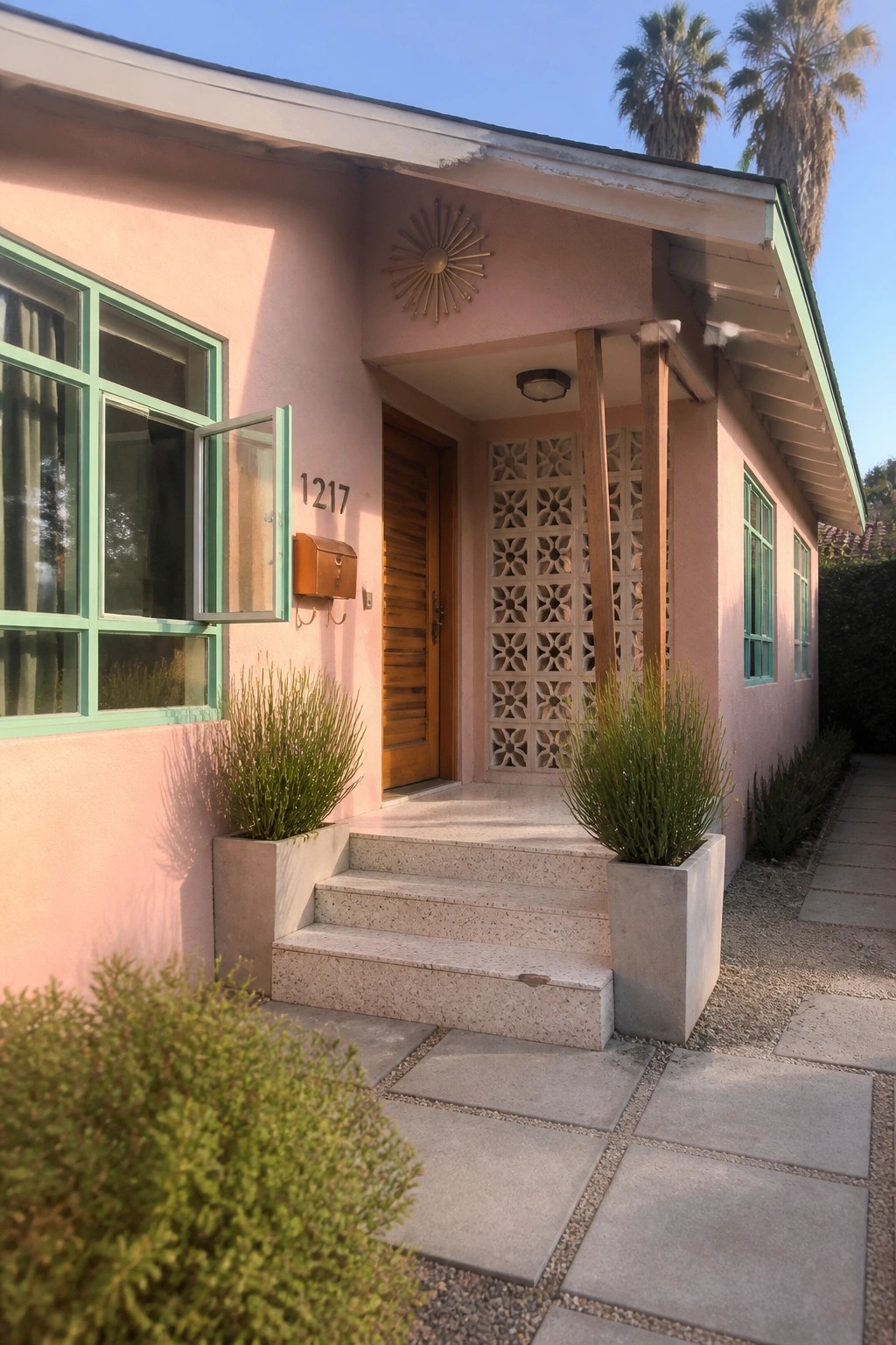 Front view of a pink stucco bungalow with mint green window frames, a wooden door, sunburst medallion above the entrance, and decorative concrete screens beside the steps.