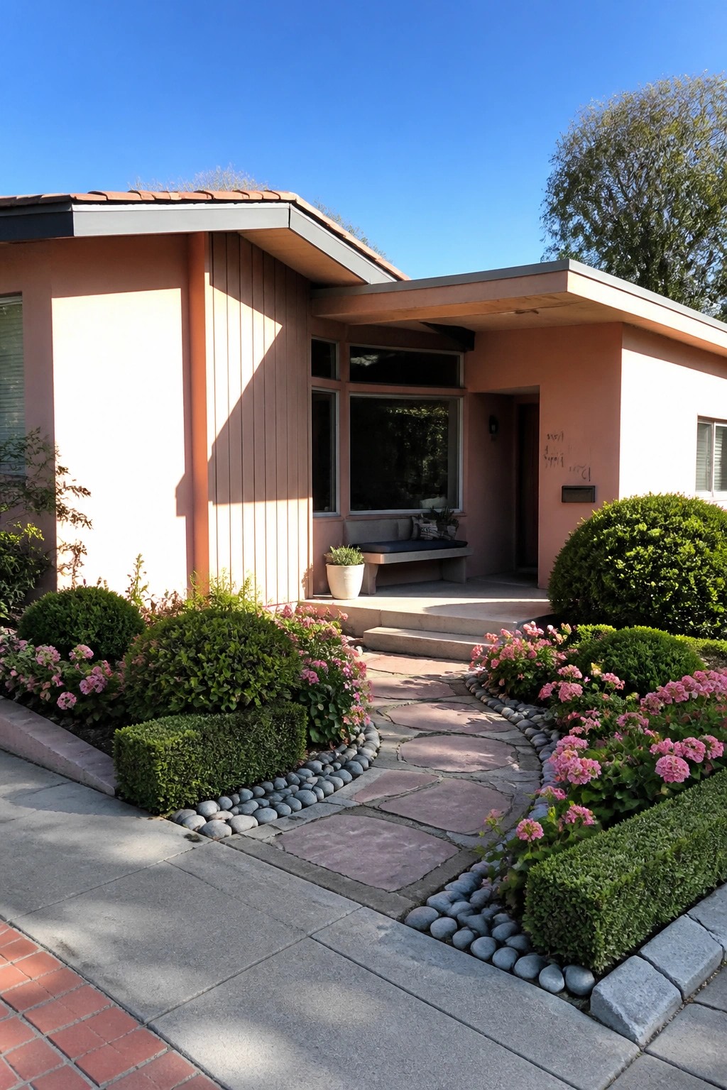 A single-story midcentury house painted in a soft peach-pink with vertical wood siding, a curved stone path bordered by pebbles and flowering shrubs, and a small covered entry.