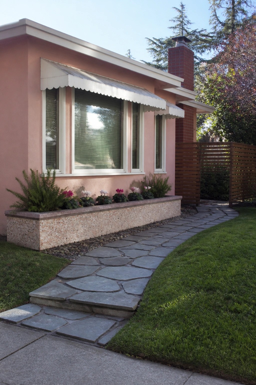 Pink house with white awning, flower bed, stone path, and wooden fence.