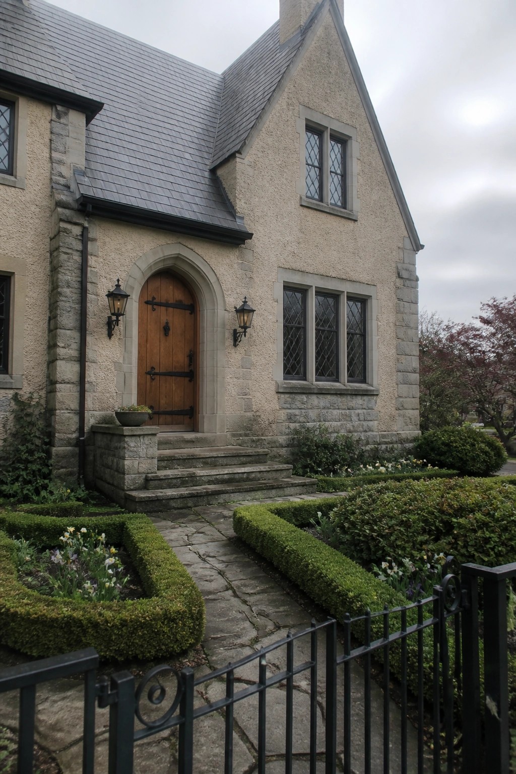 A beige stone house with a gray slate roof has an arched wooden front door framed by stone, with leaded windows and a stone path leading to the entry.