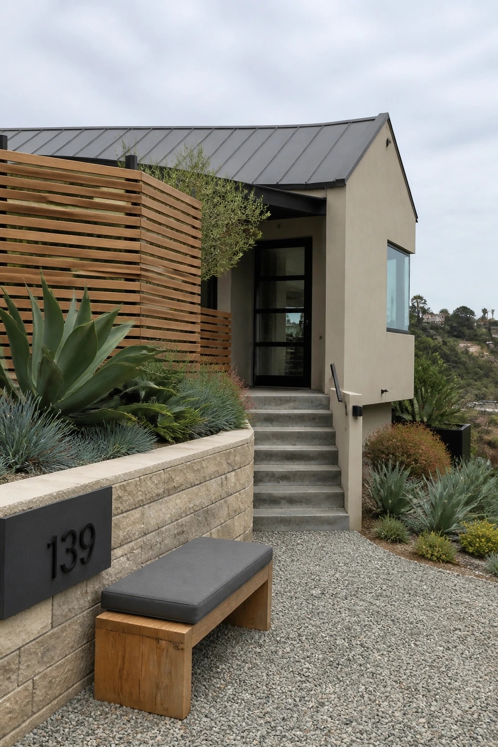 Modern beige house exterior with gray metal roof, wooden privacy screen, stone wall, and concrete steps leading to a glass door.