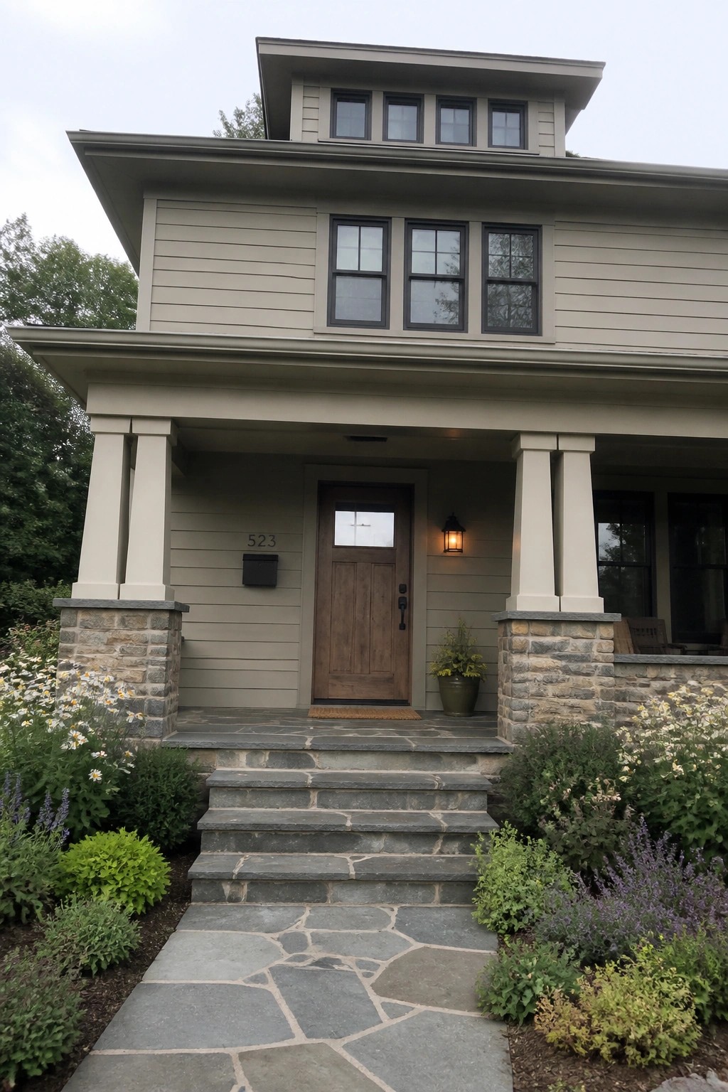 Two-story taupe house with wooden door, stone porch pillars, steps, and flower garden.