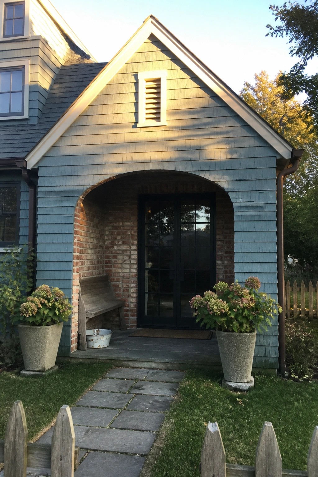 A teal shingled house with an arched brick entry surrounding black double doors, a stone path in front, and potted plants on either side.
