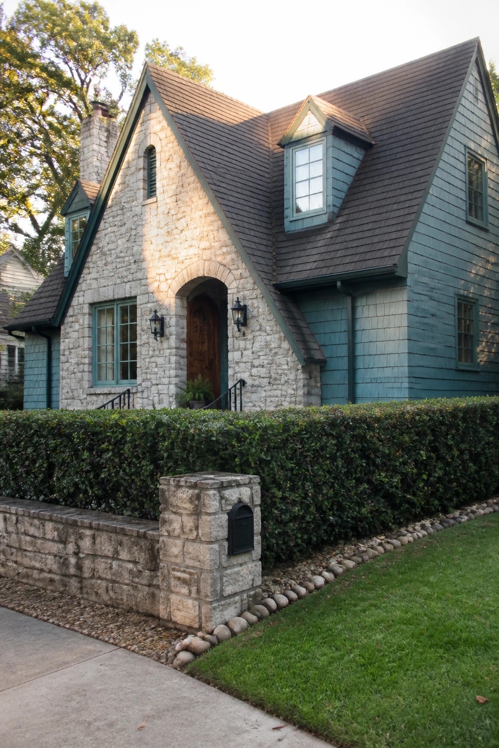 A two-story house with light stone on the left side and dusty teal wood siding on the right, a dark shingled roof, an arched wooden door, and a tall hedge in front.
