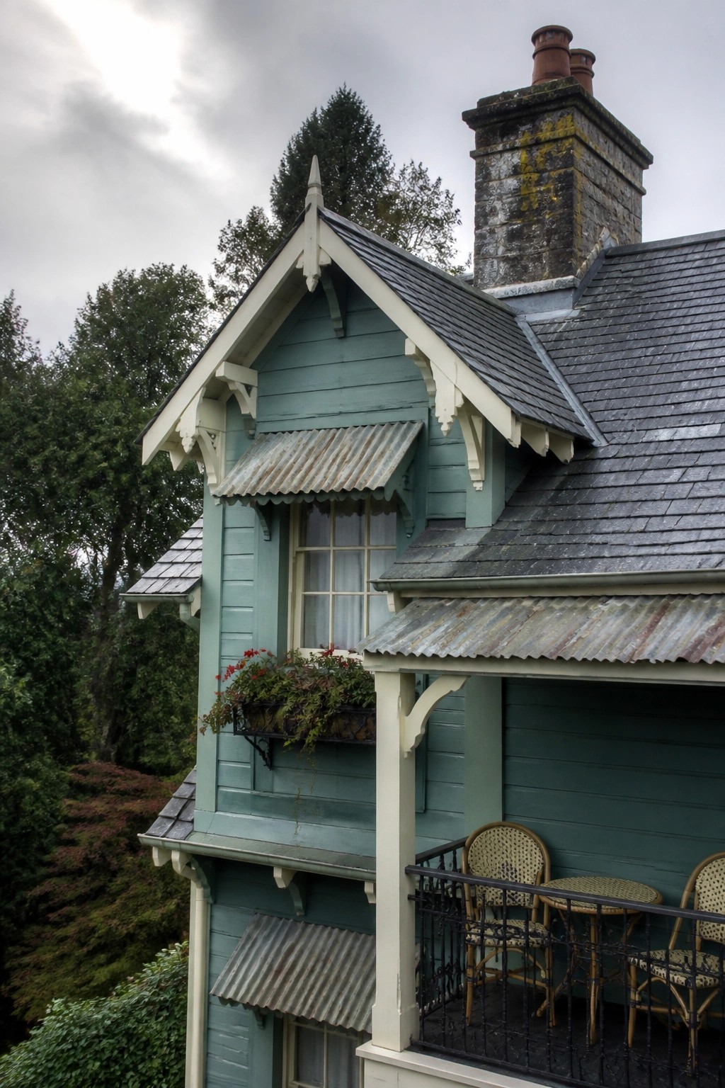 A multi-story house with dusty teal wooden siding, white trim, a balcony with wicker chairs, metal awnings, and a stone chimney.