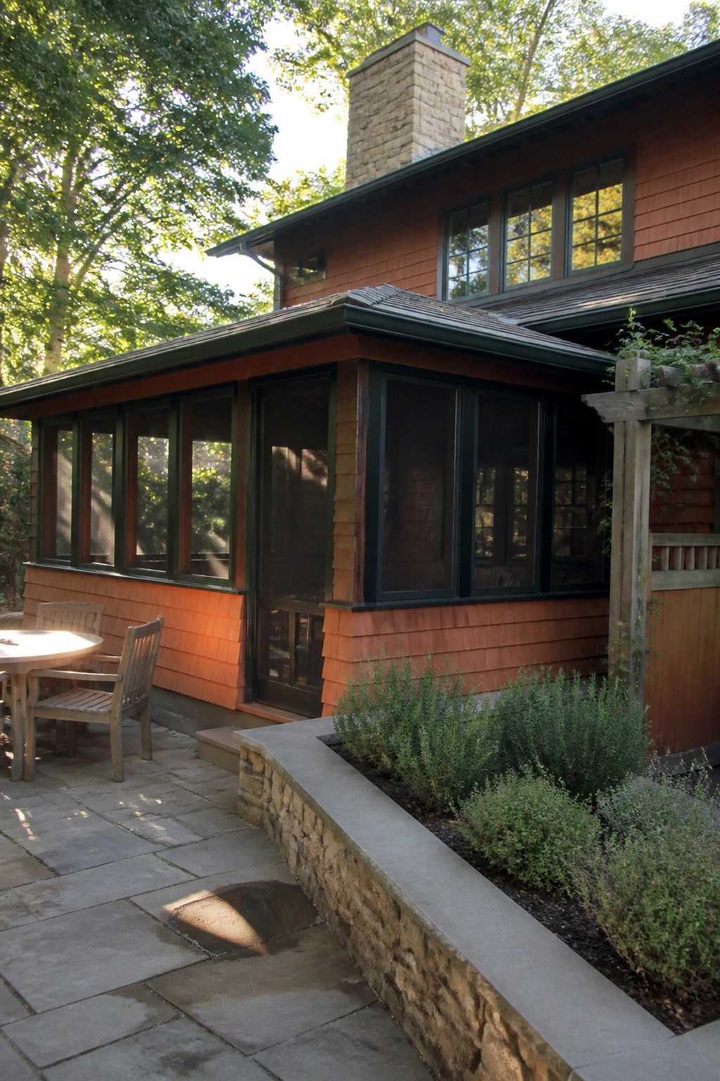 A Craftsman house exterior with orange shingle siding, a screened porch, stone chimney, patio with wooden table and chairs, and a stone-bordered garden bed.