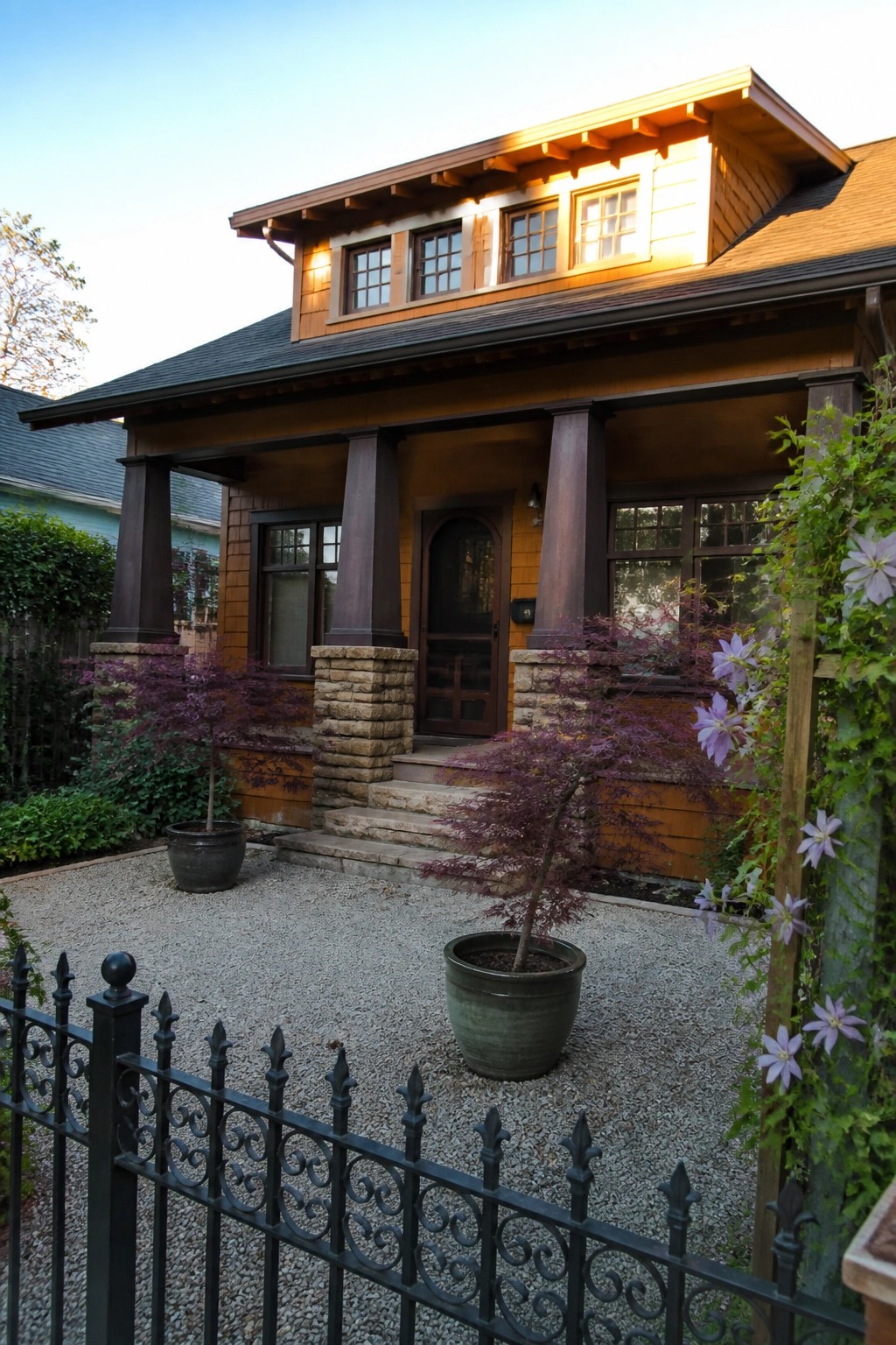 A Craftsman house with orange shingle siding, a front porch with dark columns on stone bases, concrete steps, and gravel landscaping in the foreground.