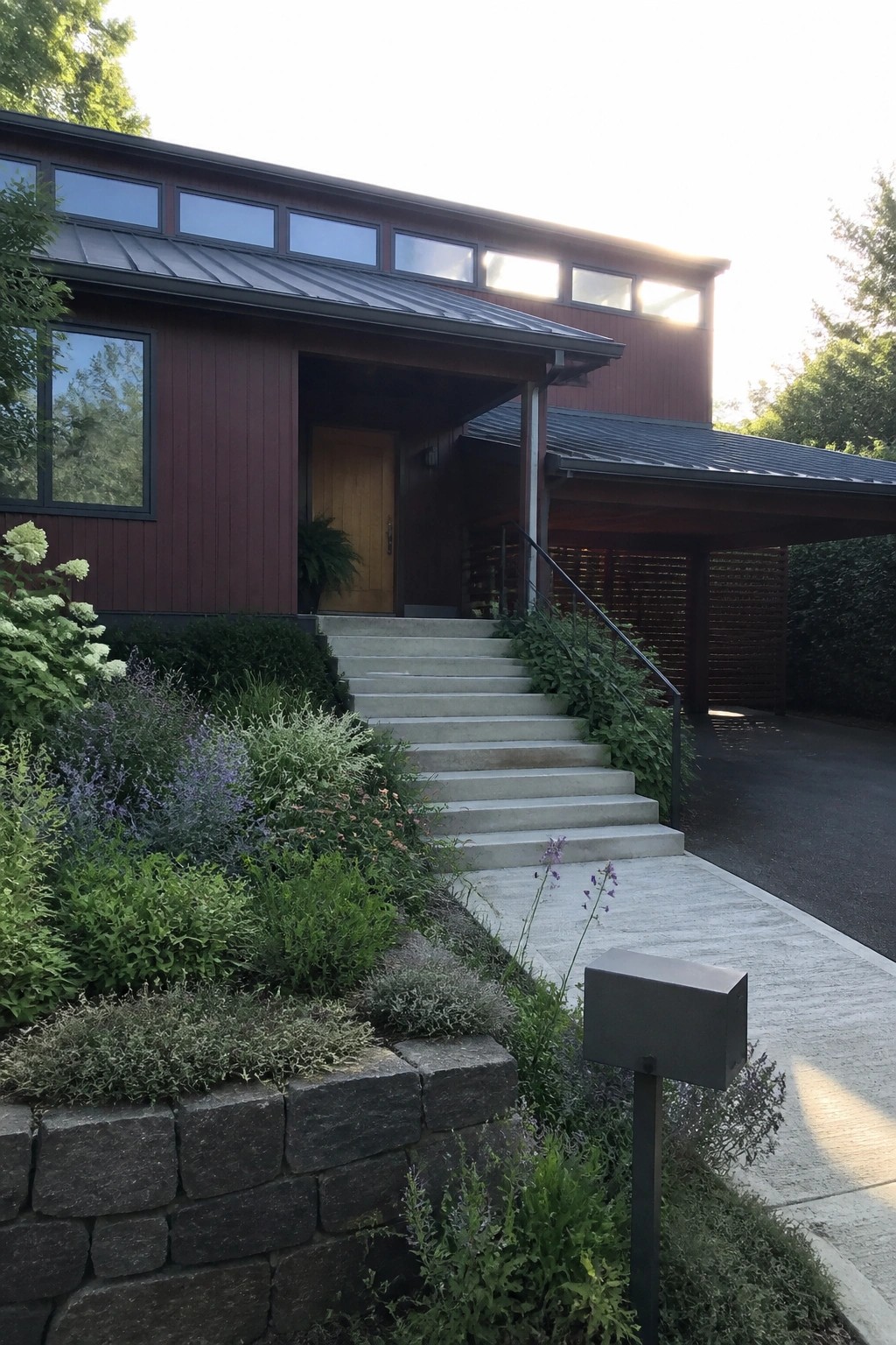 A two-story house with dark maroon wood siding, a charcoal metal roof, concrete stairs leading to a light wood door, and a stone retaining wall with landscaping in the foreground.