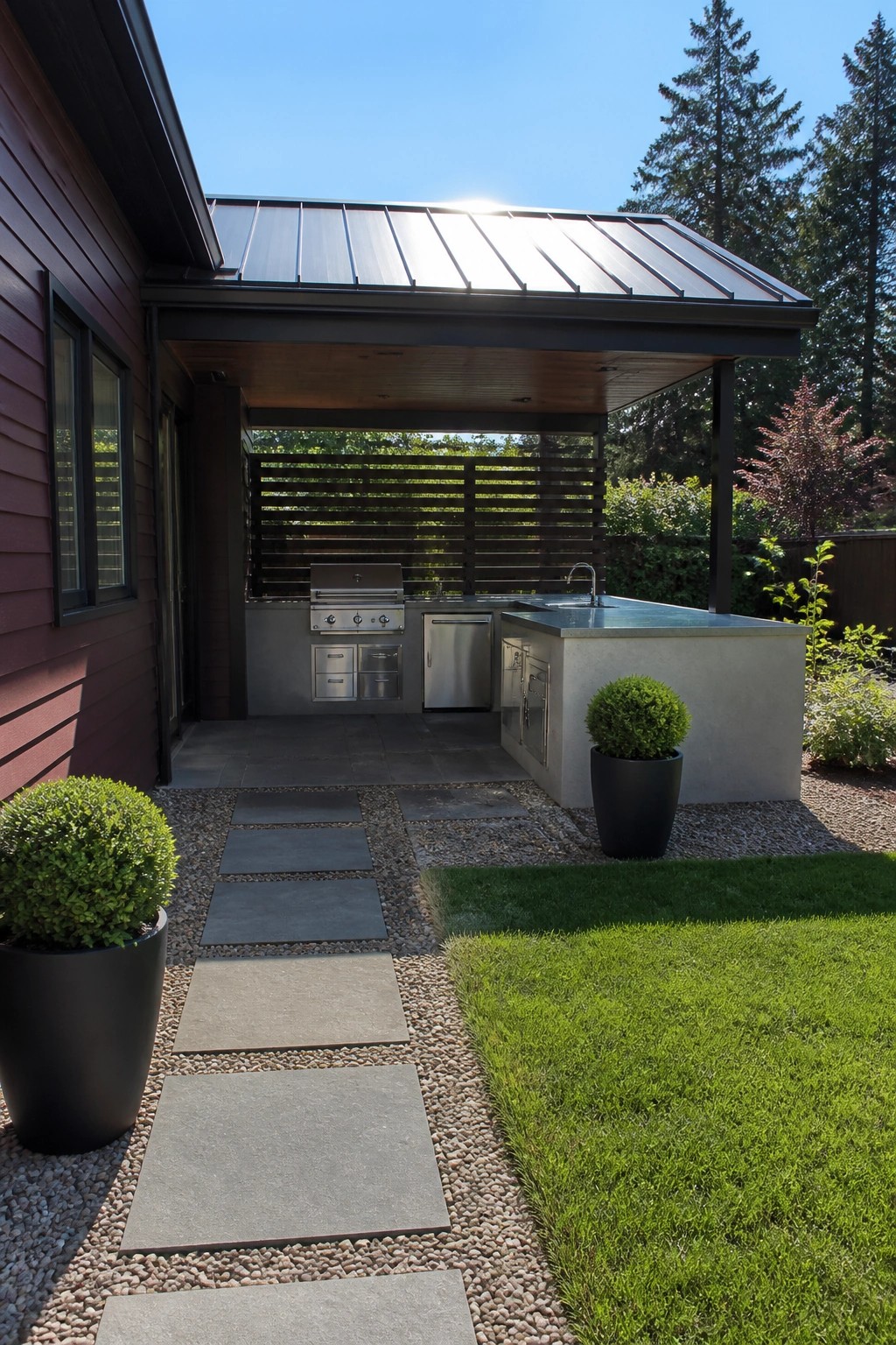 A dark maroon house exterior with charcoal metal roofing extended over a concrete outdoor kitchen that includes a stainless steel grill, sink, and cabinets, with a stone pathway and potted shrubs in the foreground.