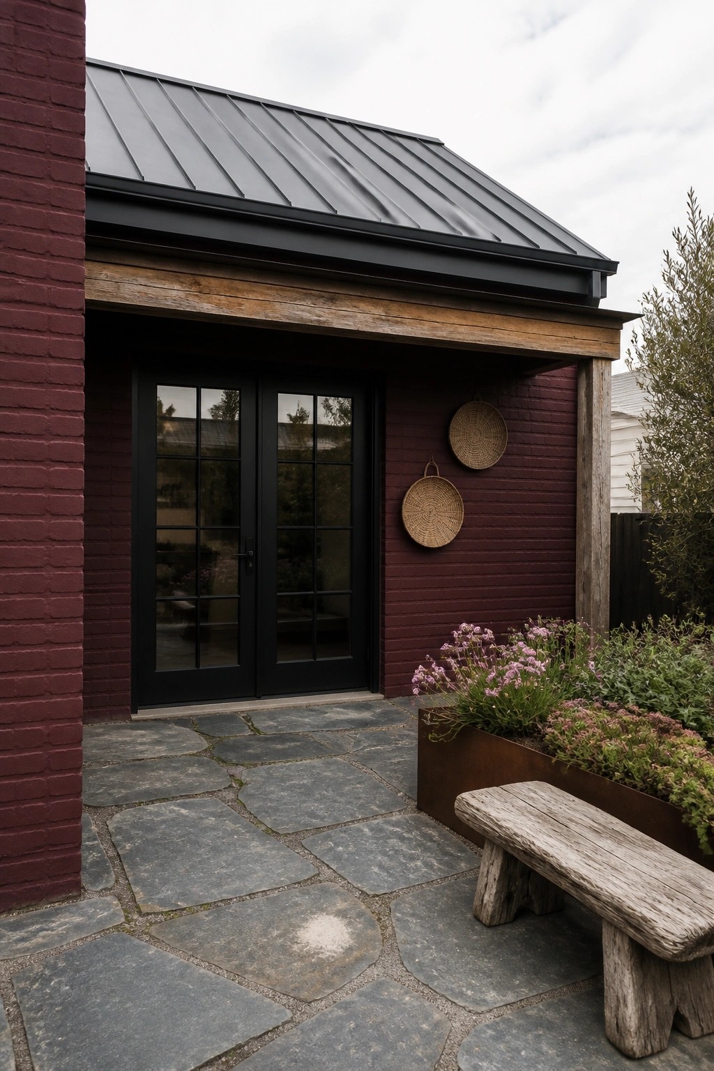 Maroon brick building with black double doors, stone patio, wooden bench, and flowers.