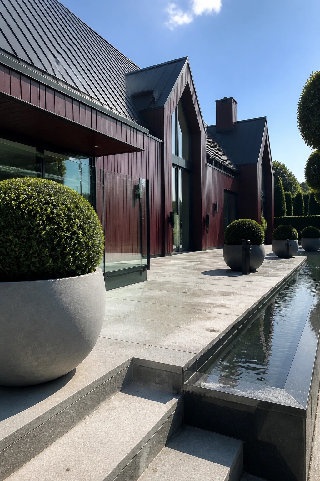 A modern house exterior with dark maroon vertical siding, a charcoal metal roof, large glass doors, and concrete planters beside a paved patio and water feature.