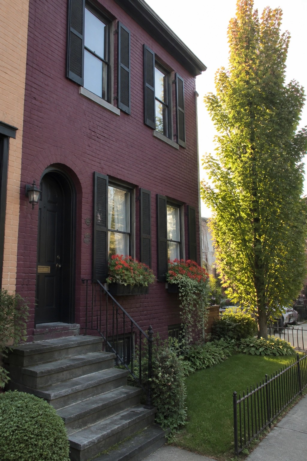 A three-story burgundy brick house with black shutters, a black arched door, concrete steps, and red flower boxes under the windows.