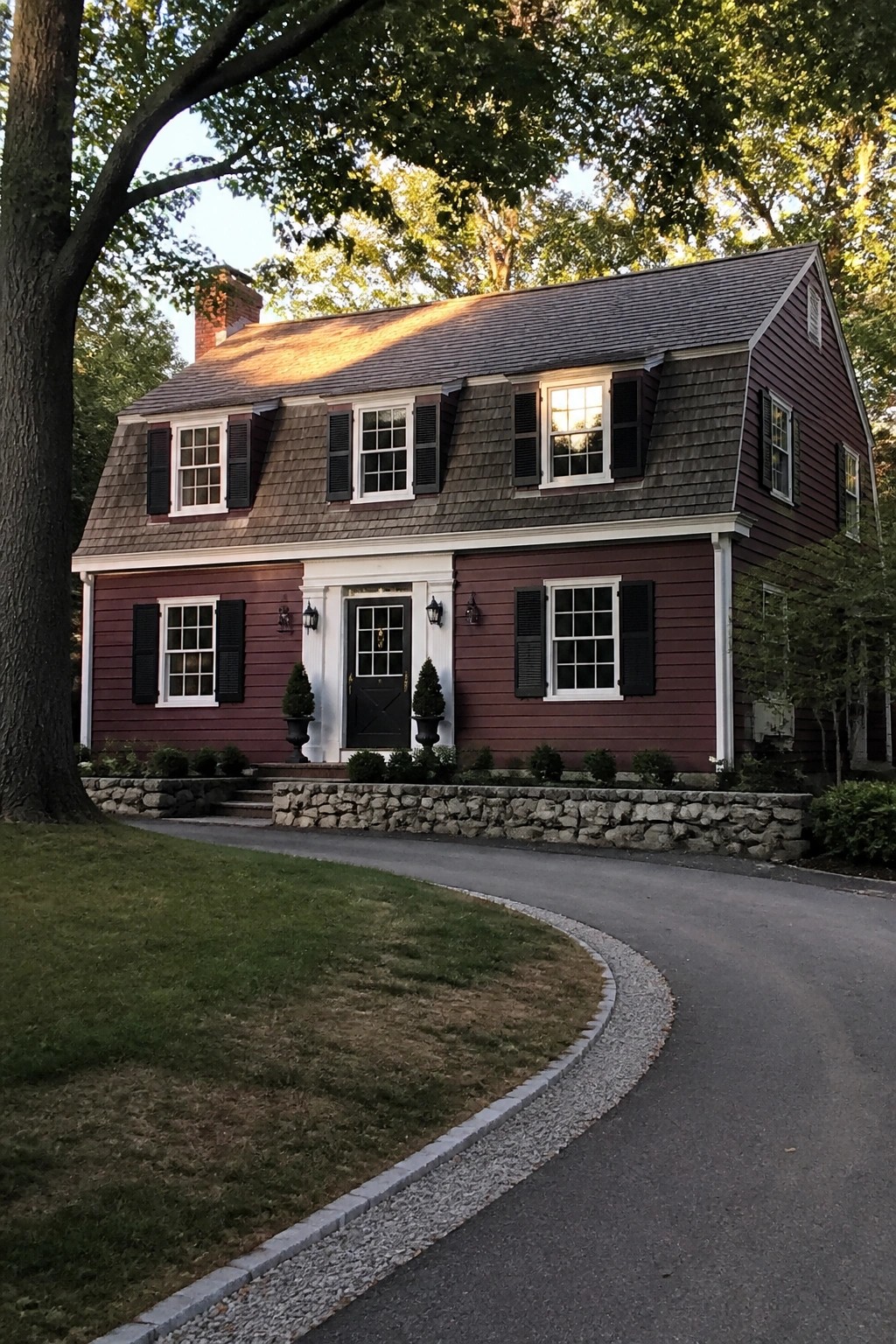 Two-story red house with black shutters, stone wall, and curved driveway