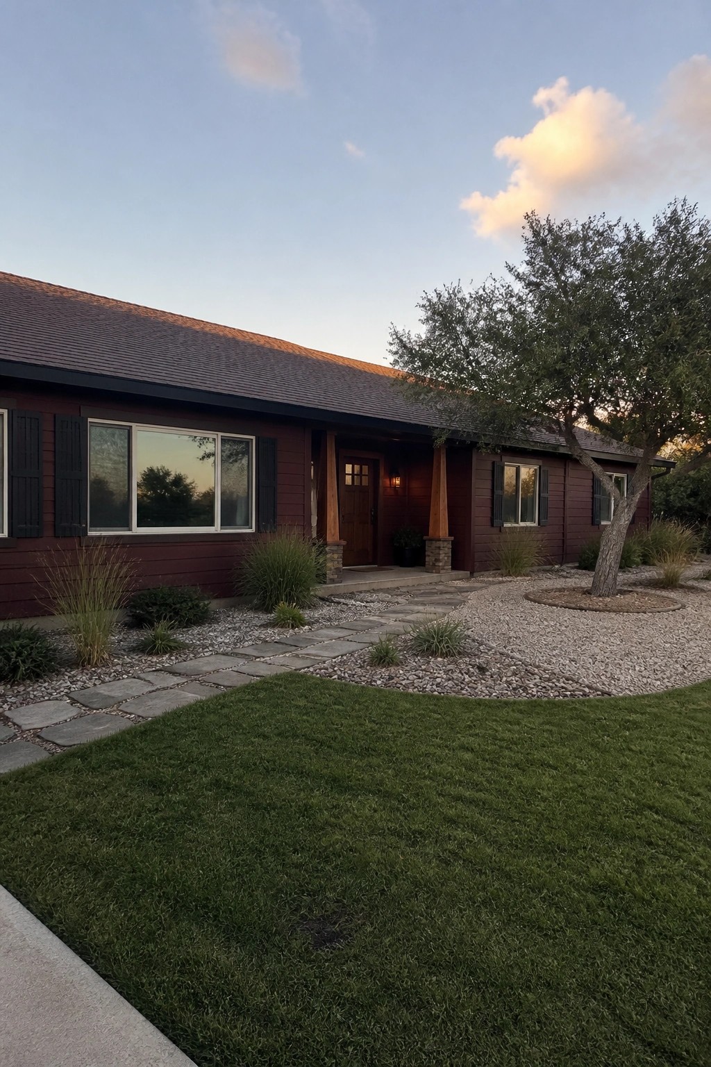 A single-story burgundy house with black shutters, a covered entry with wood posts, and a stone path curving through gravel landscaping.