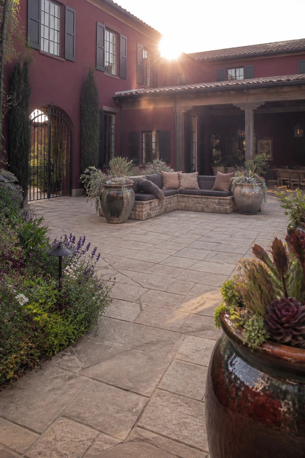 A paved courtyard beside a deep red house with black shutters, showing an L-shaped built-in bench with cushions and two large ceramic planters at each end.