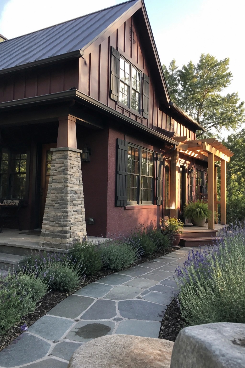 A two-story house with deep burgundy siding, black shutters, a stone porch column, and a metal roof, viewed from a stone pathway with lavender plants in the foreground.