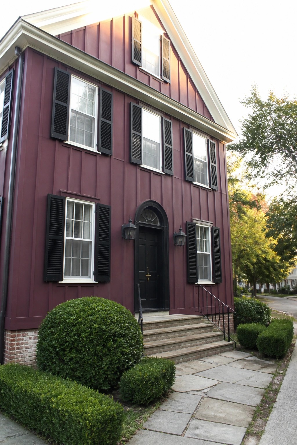 A two-story house with deep burgundy siding, black shutters on multiple windows, a black front door under an arched transom, and stone steps with black railings.