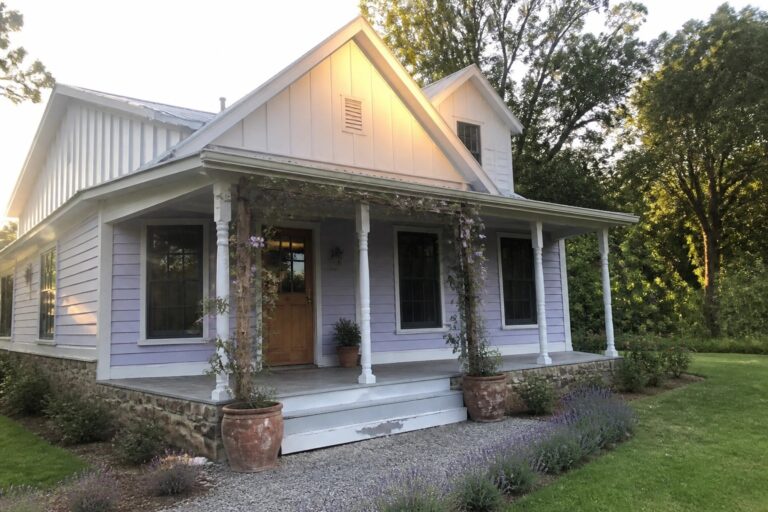 18 Fresh Light Purple House Exteriors Featuring White Board-and-Batten Siding A light purple house with white trim, a wooden front door, black-framed windows, and climbing vines on the porch structure.