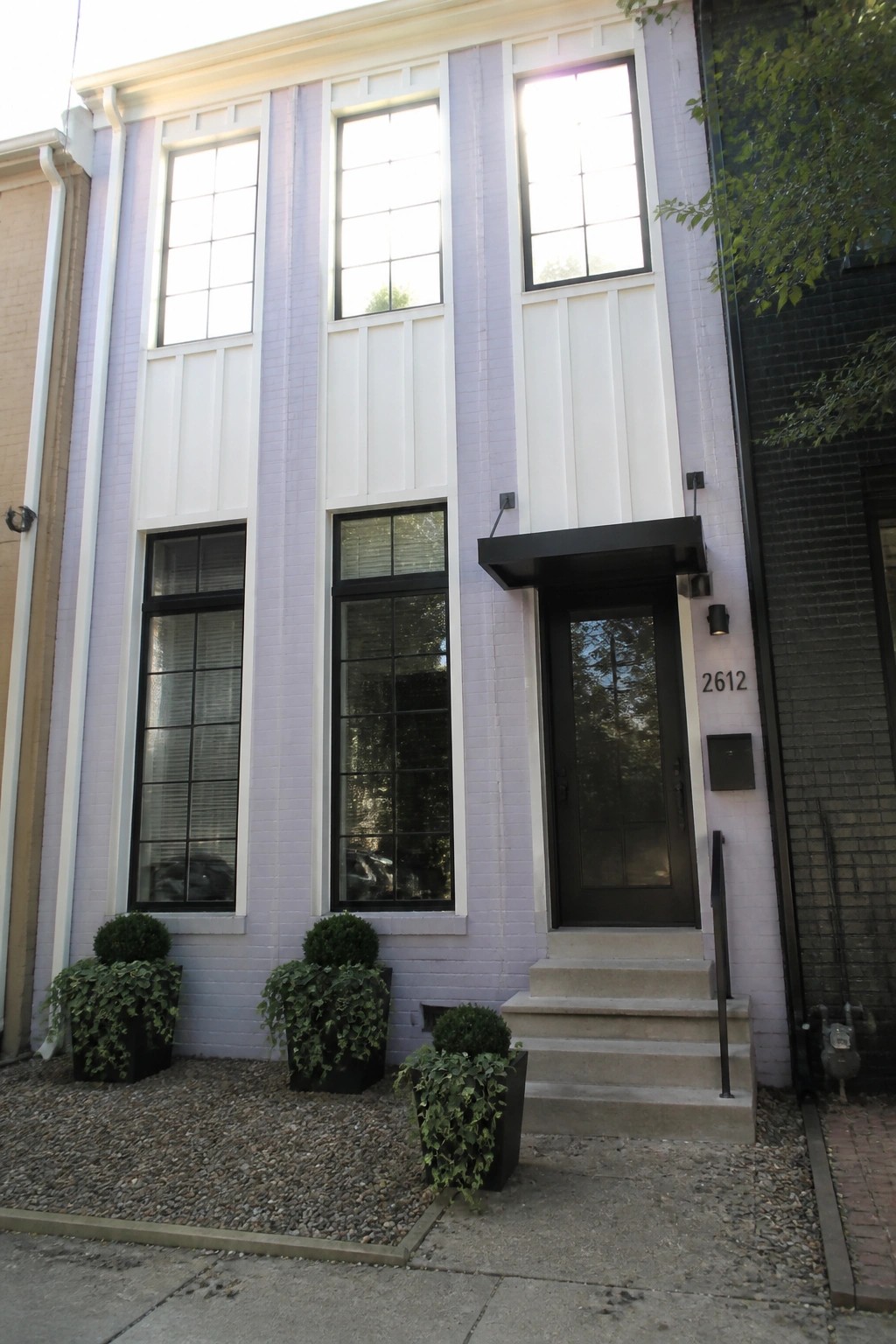 A narrow light purple brick house exterior with white board-and-batten panels, black-framed windows, and a black front door with steps.