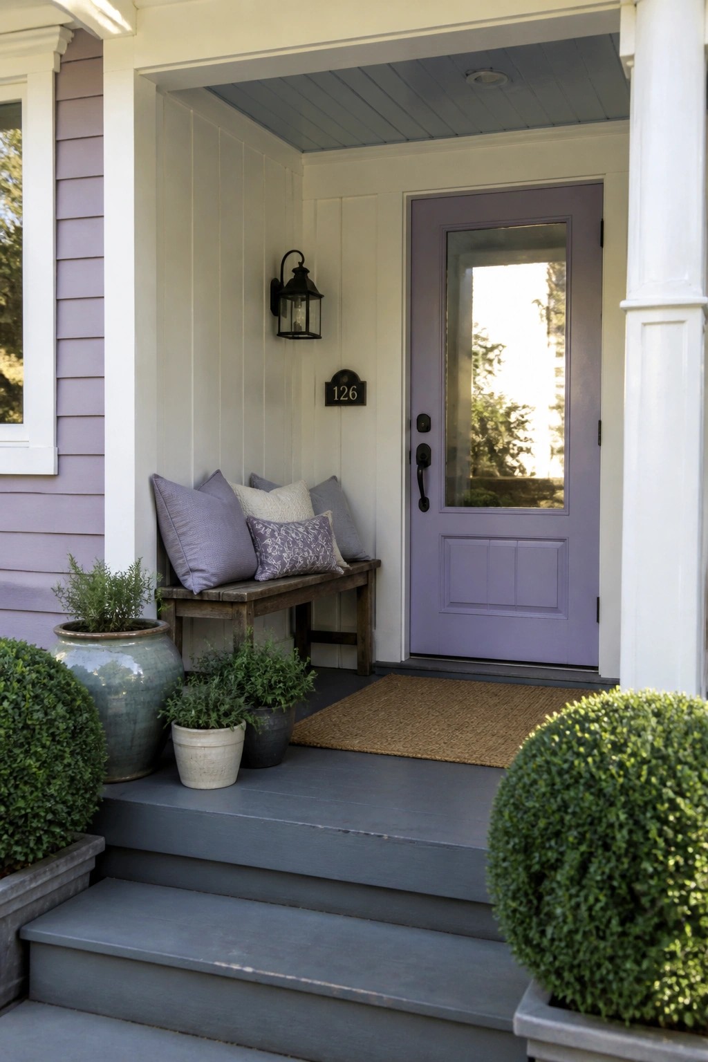 A front porch with light purple siding on the left, white board-and-batten walls, a matching purple door, a wooden bench with pillows, and potted plants beside gray steps.
