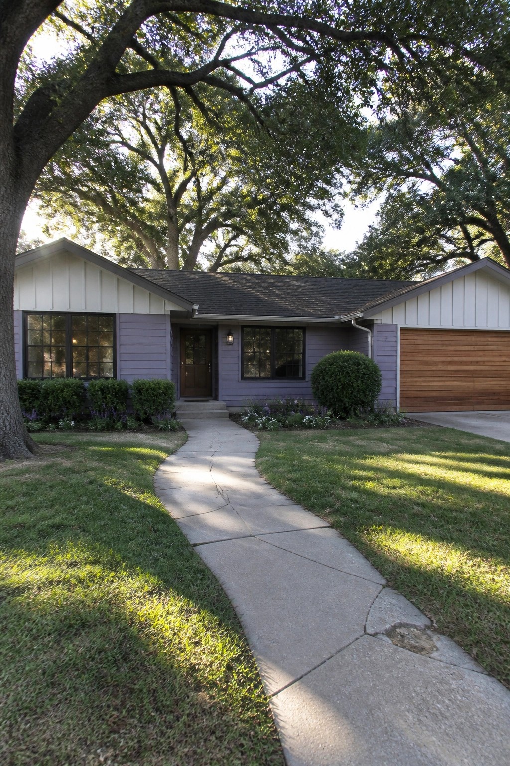 A single-story house with light purple siding, white board-and-batten on the gables, a brown front door, and a wood garage door set among large trees and a concrete walkway.