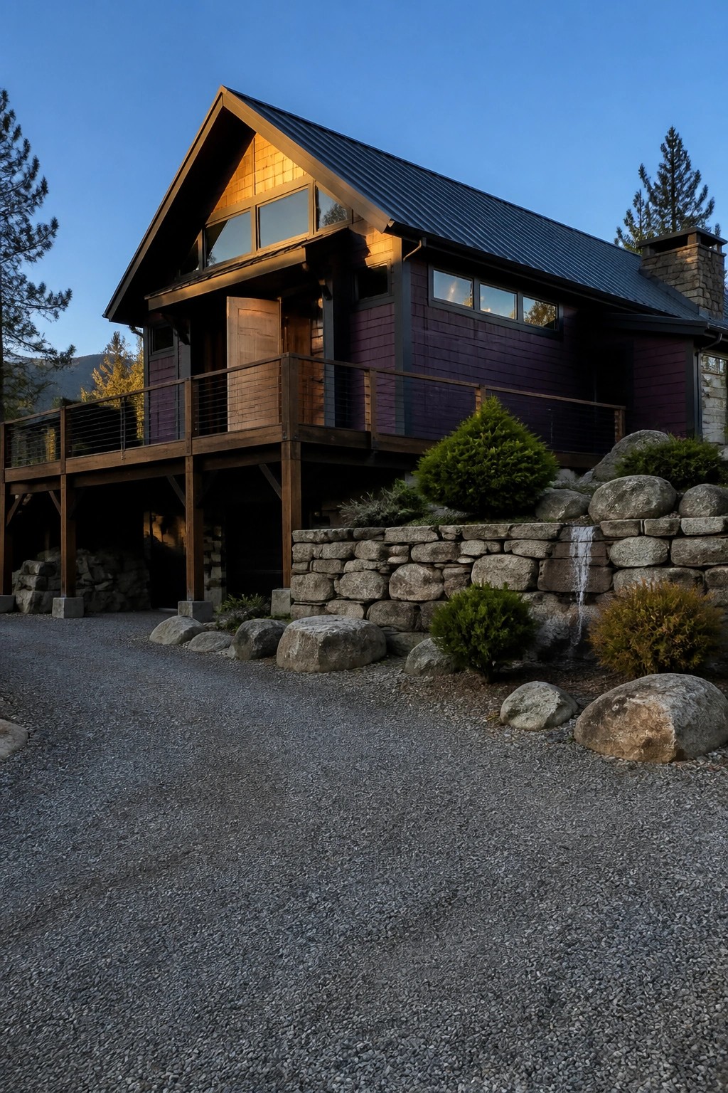 Modern purple wooden cabin with balcony, stone wall, and rocky landscaping at dusk