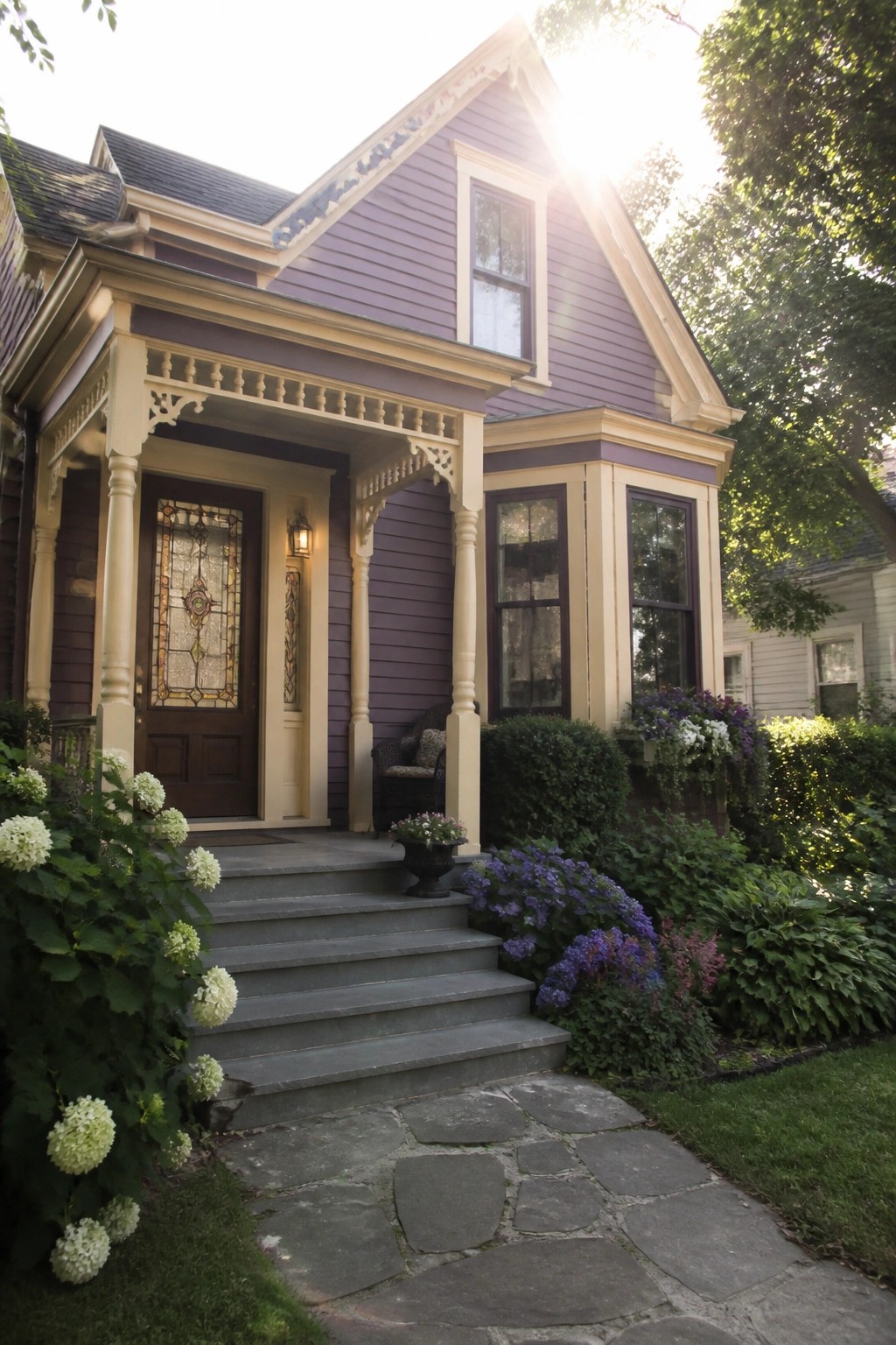 A two-story house with purple horizontal lap siding, cream trim, a covered front porch, stone steps, and a stone pathway bordered by green shrubs and white and purple flowers.