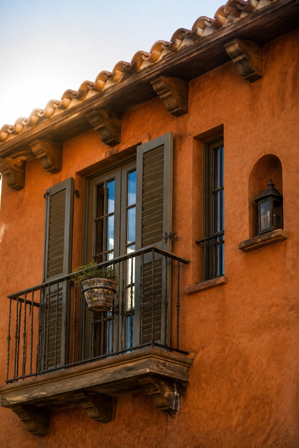 Orange stucco wall with balcony, potted plant, shutters, and lantern niche.