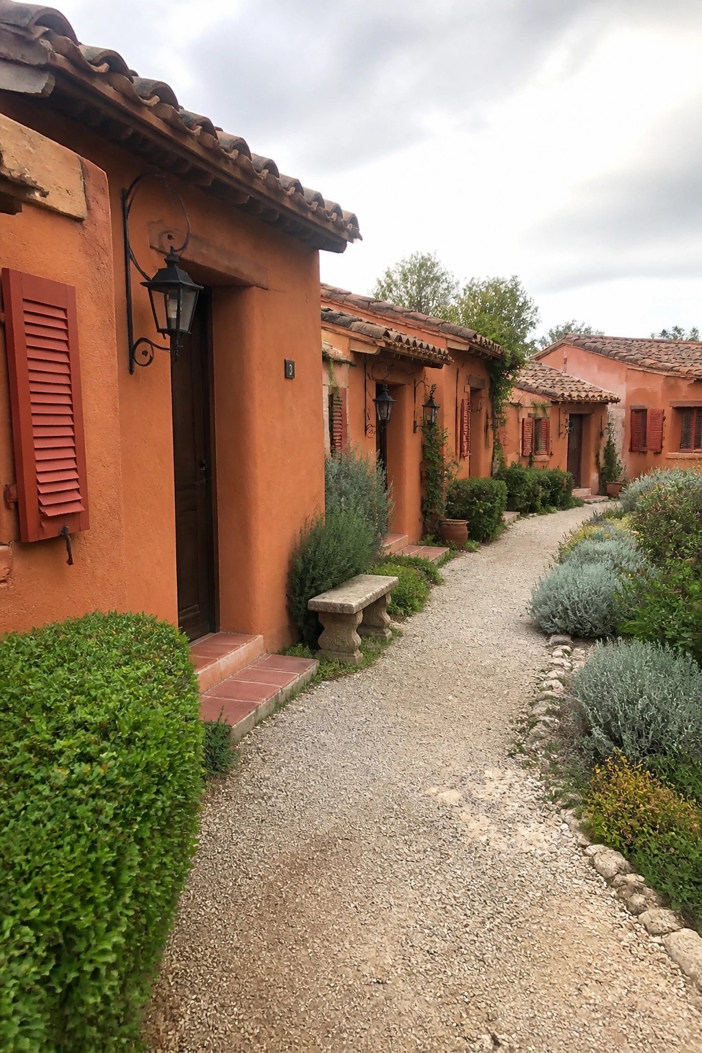 A row of orange stucco buildings with terracotta tile roofs along a gravel path, with wooden doors, red shutters, and wall-mounted lanterns.