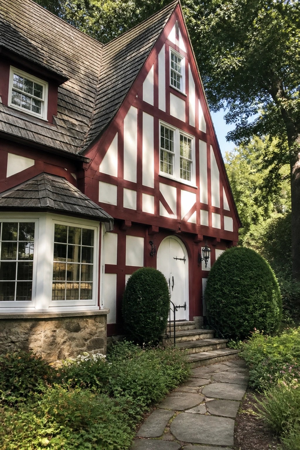 A two-story Tudor-style house exterior with burgundy half-timbering over white panels, a white arched door, stone foundation, and surrounding shrubs with a stone path.