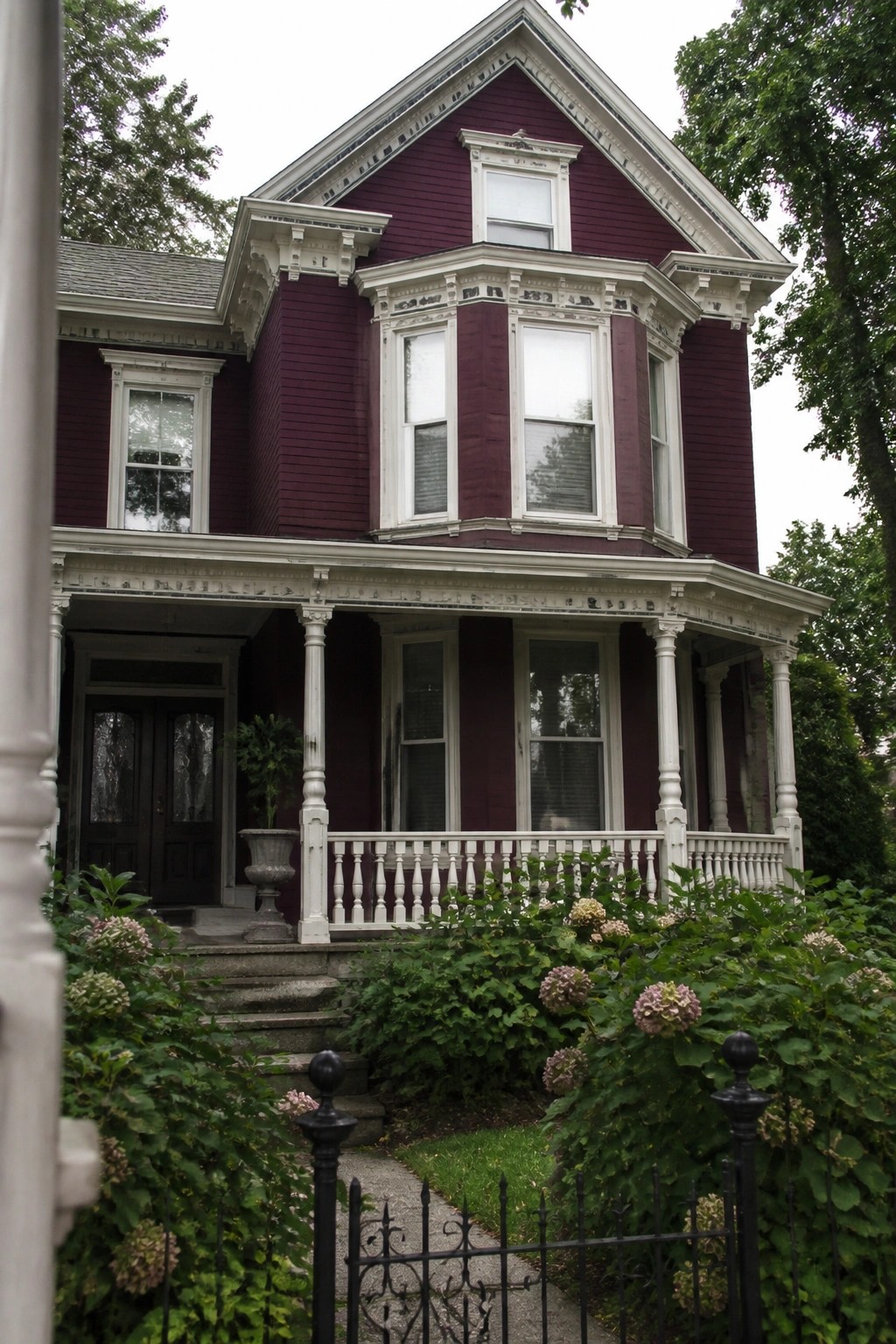 A two-story burgundy house with white trim around windows, porch columns, and eaves, plus a front garden with hydrangeas and a black fence.