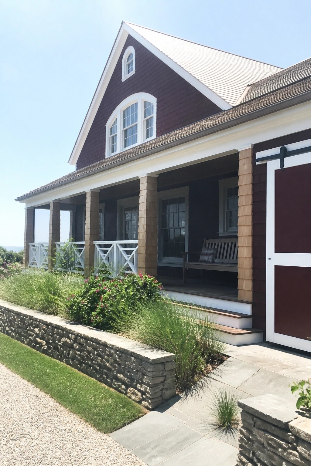 A two-story house with burgundy shingle siding, white trim on windows and porch, a covered entry with wooden columns, and a stone retaining wall along the front.