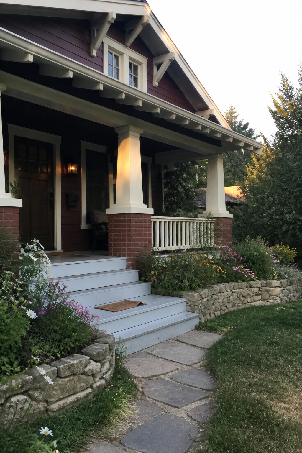 A burgundy house exterior with white trim, a covered porch supported by white columns on brick bases, gray painted steps, and a stone pathway bordered by flower beds.