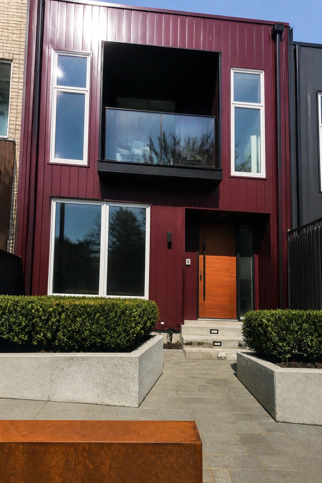 A modern two-story house with deep burgundy siding, white-framed windows of varying sizes, a wood front door, and concrete planters along the walkway.