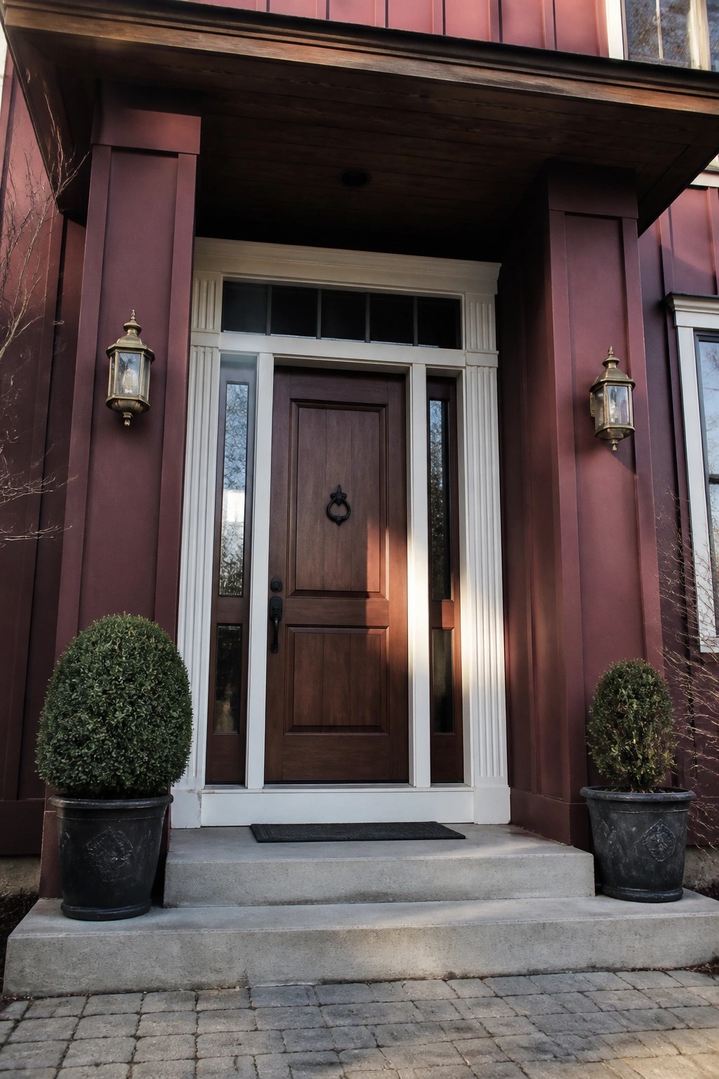 Front entrance of a house with maroon vertical board and batten siding, a dark wood door with white trim, two wall lanterns, and potted shrubs on concrete steps.