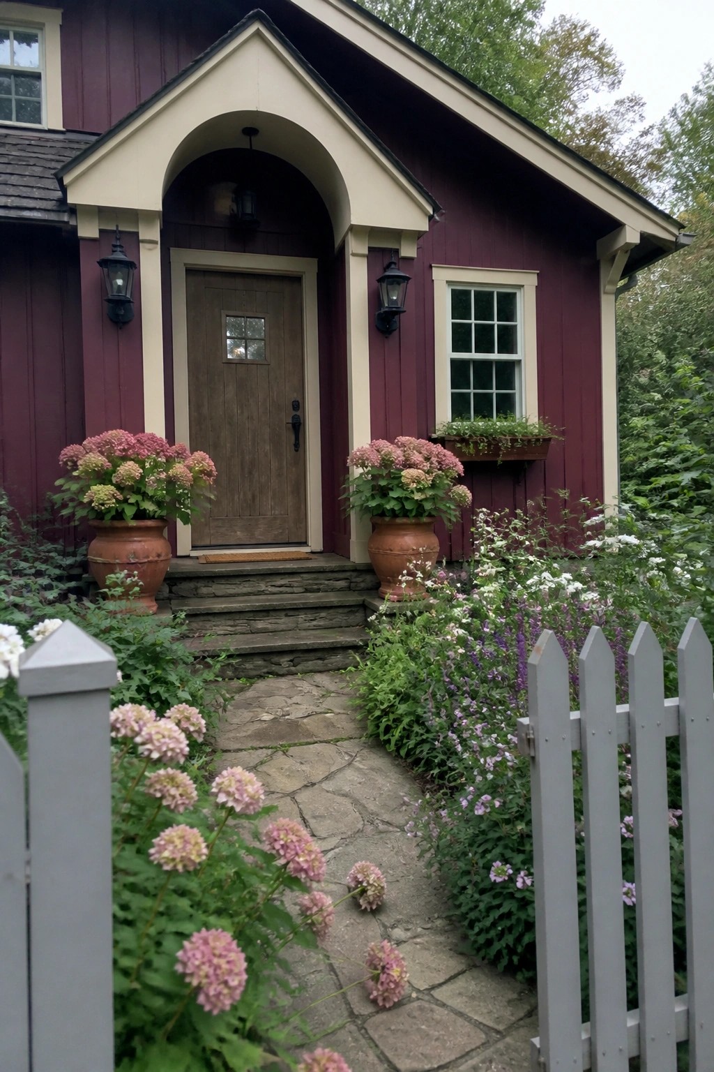 A house exterior with maroon vertical board and batten siding, an arched entry, a wooden door, and two large potted plants on stone steps.