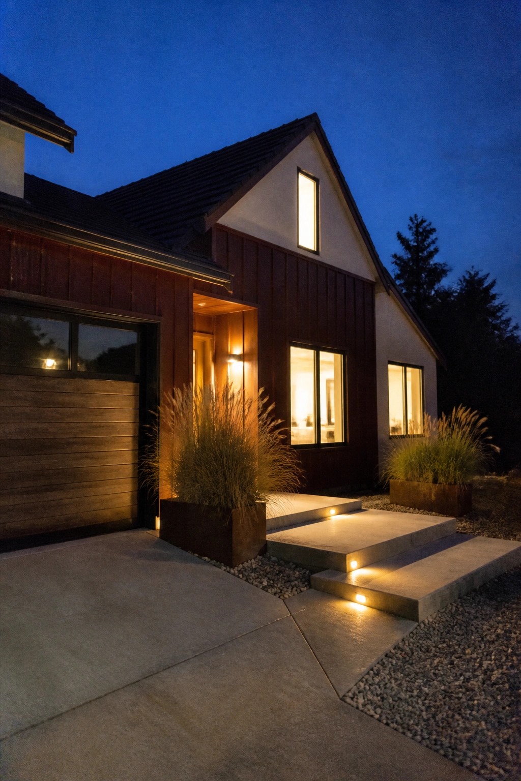 Modern house with dark red siding, lit windows, concrete steps, and tall grass planters at dusk.