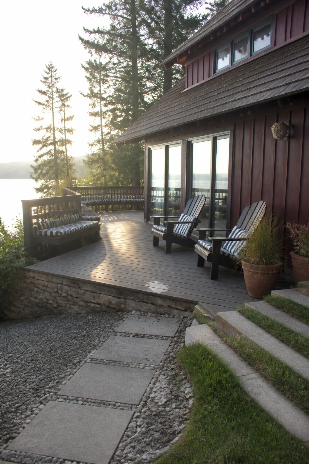 House exterior with vertical maroon board and batten siding, large glass doors, and a wooden deck with chairs overlooking a lake.