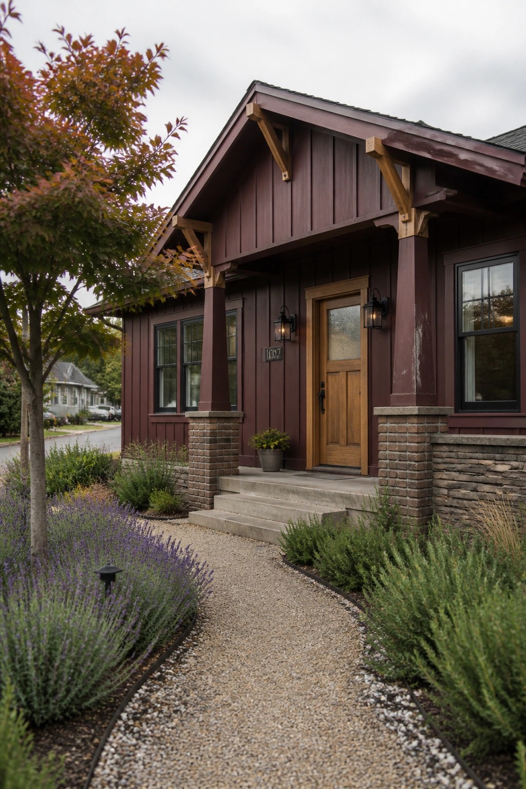 Front view of a house exterior with dark maroon vertical board and batten siding, wooden front door, stone porch pillars, and gravel pathway.