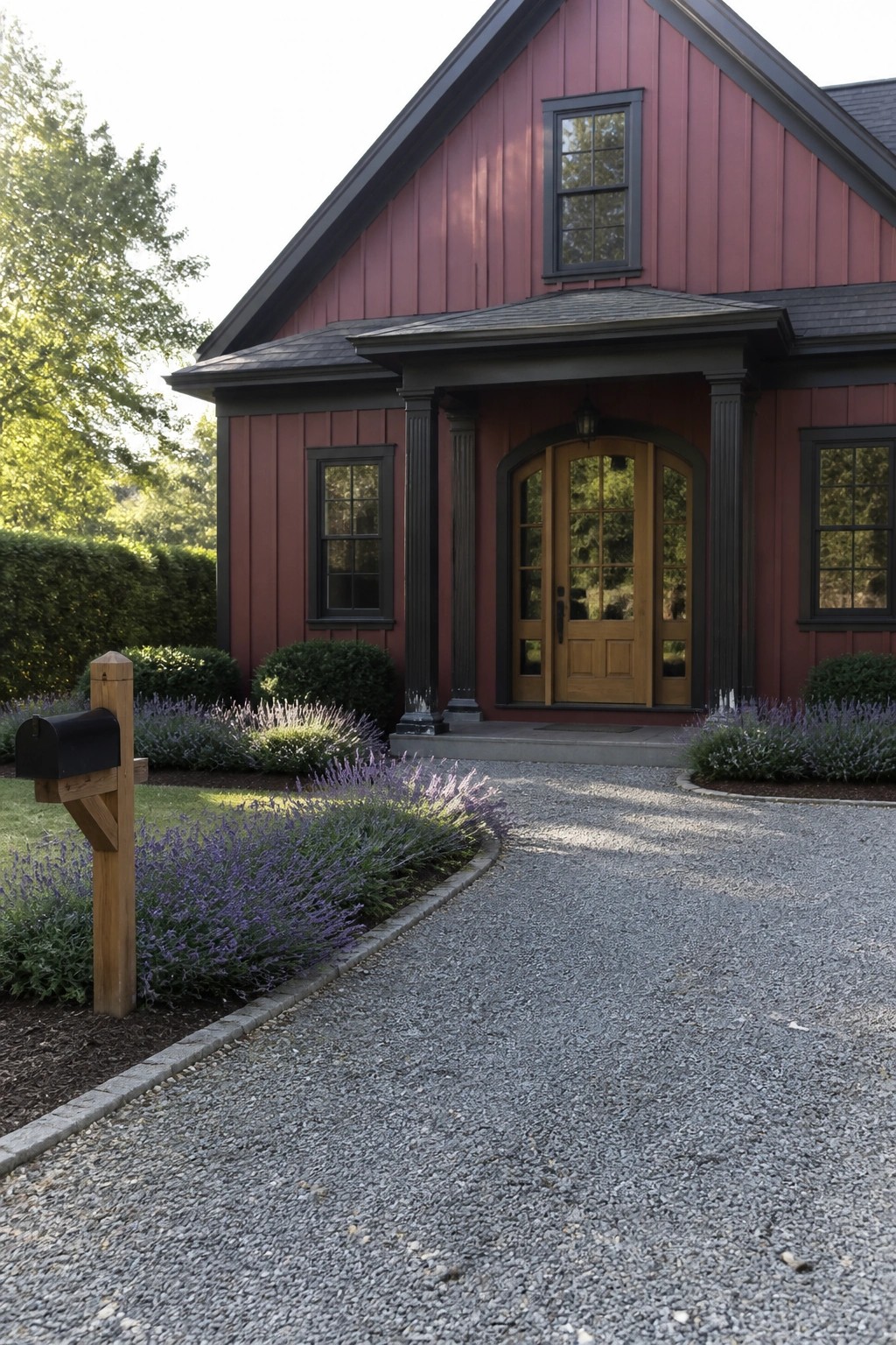 A house with deep maroon vertical board and batten siding, dark trim around windows and columns, a wooden arched front door, and a gravel driveway bordered by lavender.