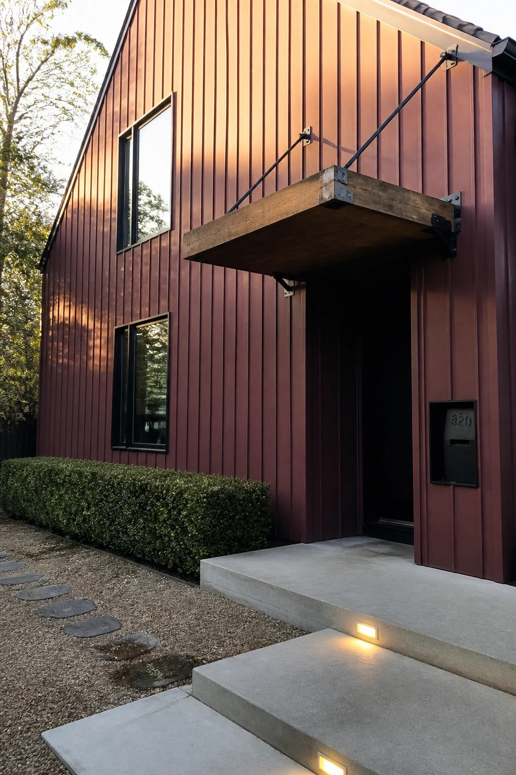 A house exterior with vertical maroon board and batten siding, a wooden entry canopy supported by metal brackets, and concrete steps with small lights.