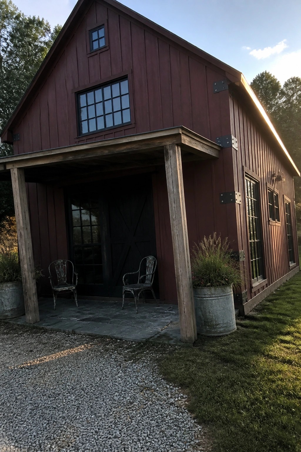 A burgundy barn-style house exterior with black-framed windows, a covered porch with two metal chairs, and large metal planters beside a gravel path.