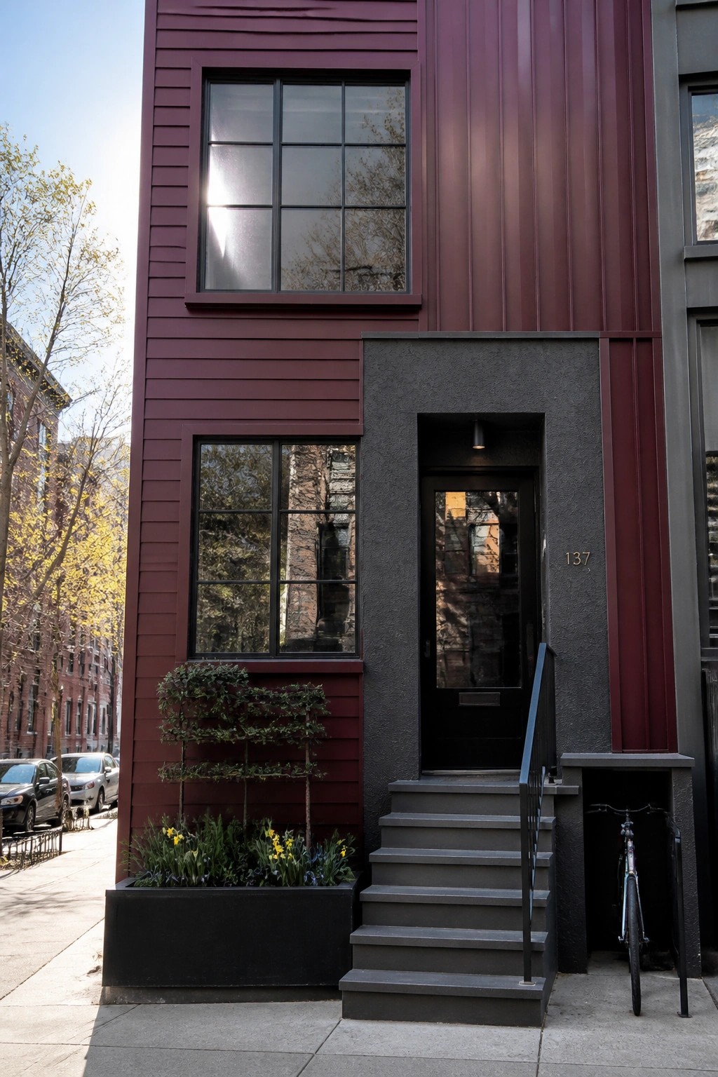 A narrow burgundy house exterior with black-framed windows, a black door, gray concrete steps, and a small front garden bed.