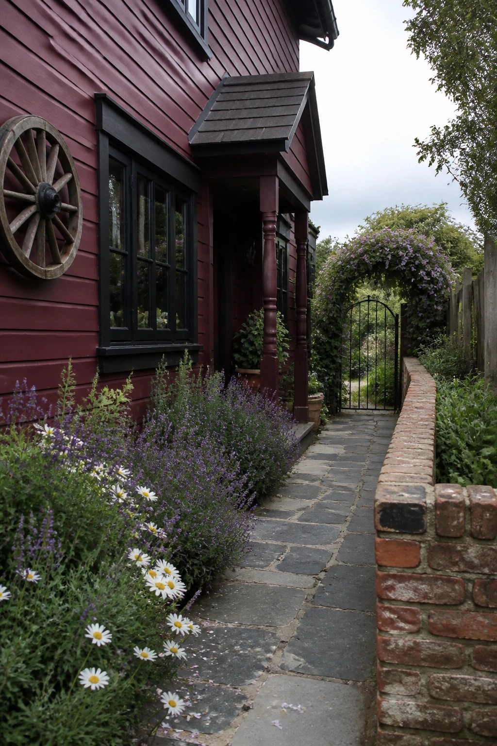 A burgundy wooden house with matte black window frames, a stone path beside a brick wall, and flowering plants along the front.