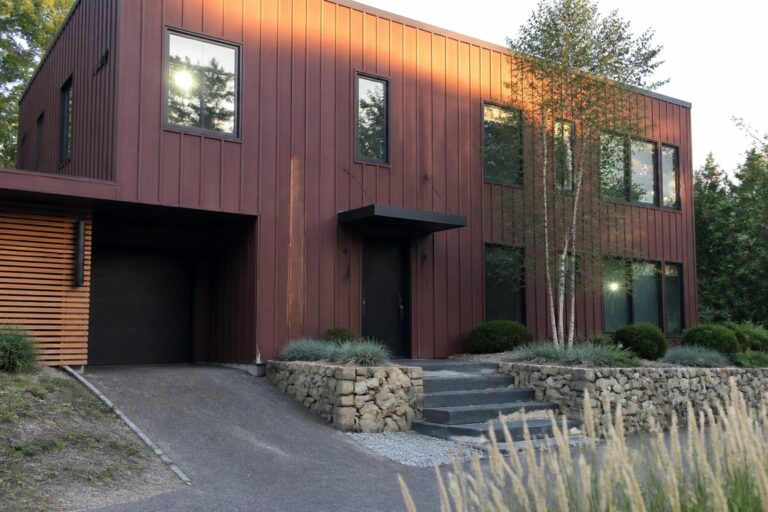A modern two-story house with vertical burgundy metal siding, matte black windows and door, and a dark garage door on the left.