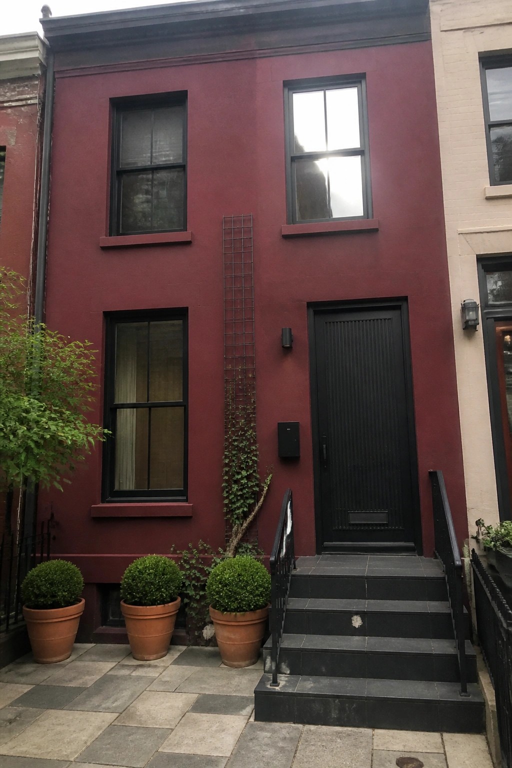 A narrow burgundy row house with matte black windows, a black door, black stairs, and three potted shrubs on a paved sidewalk.