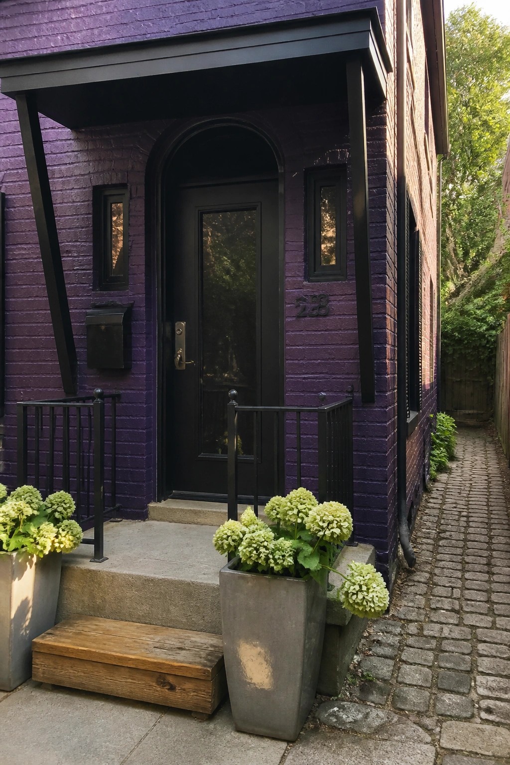 A dark purple brick house exterior with black trim, an arched black door, and two concrete planters holding green hydrangeas beside concrete steps.