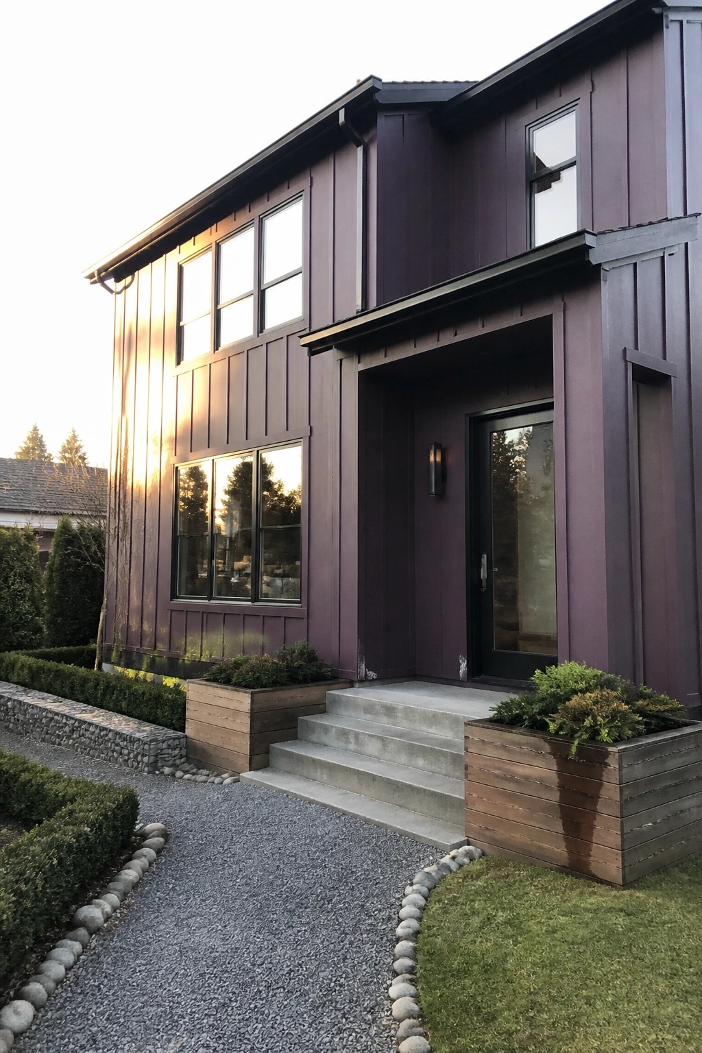 A two-story modern house with dark purple vertical siding, black-framed windows and door, concrete steps, and wooden planters flanking the entry.