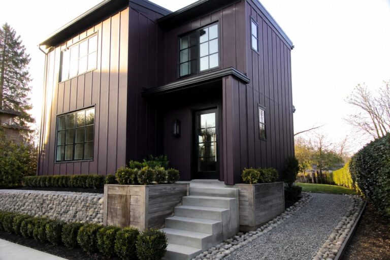 A two-story modern house with dark purple vertical siding, black-framed windows and door, concrete steps, and wooden planters flanking the entry.